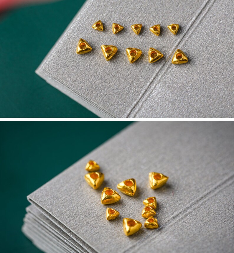May include: A collection of small, gold-colored triangle-shaped beads with a hole in the center. The beads are arranged in a pattern on a gray surface.