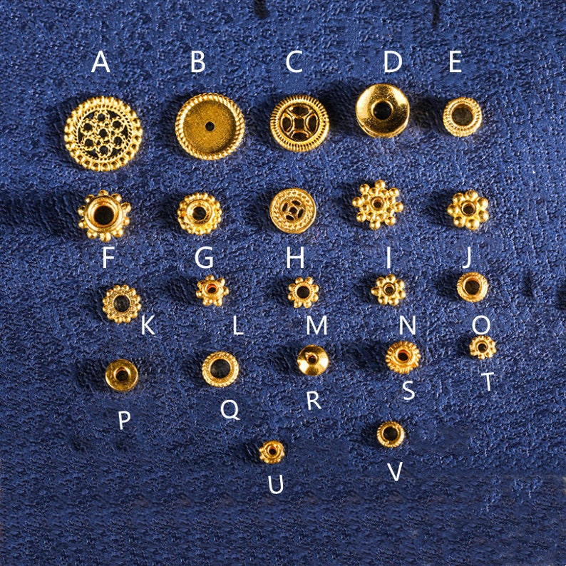 May include: Assortment of 26 gold-colored metal beads in various shapes and sizes. The beads are arranged in rows labeled with letters A through V.