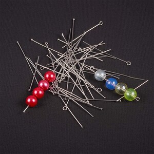 May include: A pile of silver head pins with a loop at the end, scattered on a black surface. There are four beads on the left side of the image, three red and one white. On the right side of the image, there are three beads, one light blue, one dark blue, and one green.