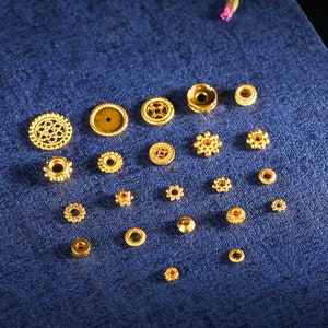 May include: Assortment of gold-colored metal beads in various shapes and sizes, including round, star-shaped, and decorative designs. The beads are arranged on a blue surface.