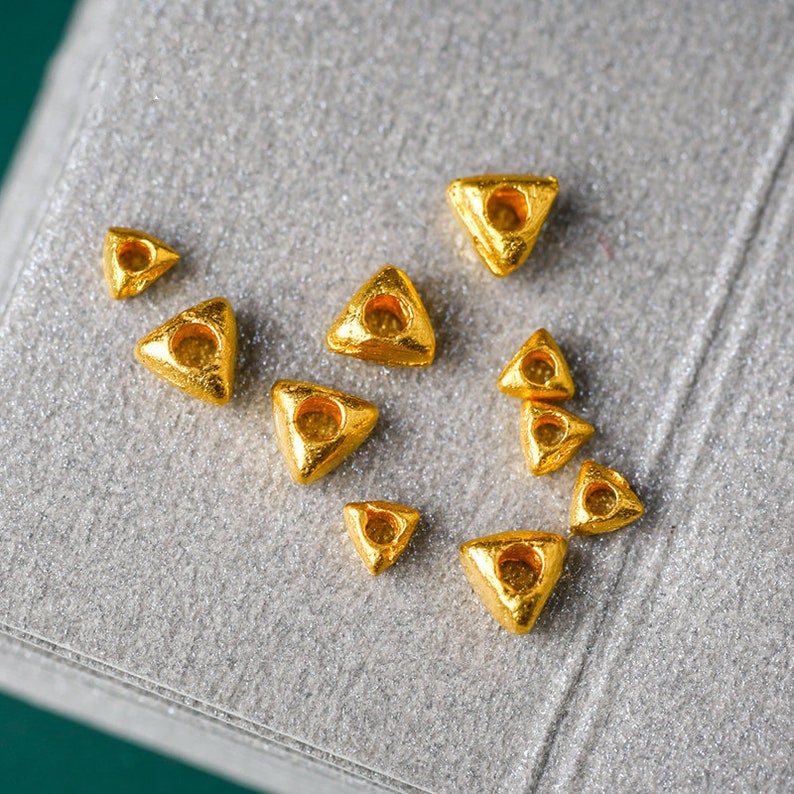 May include: A collection of small, gold-colored triangle-shaped beads with a hole in the center. The beads are scattered on a gray, textured surface.