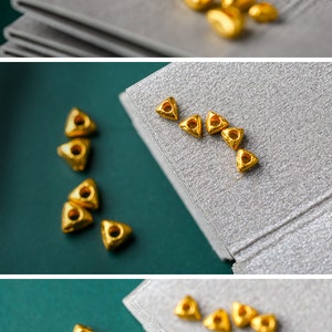 May include: A collection of small, gold-colored triangle-shaped beads with a hole in the center. The beads are scattered on a gray surface.