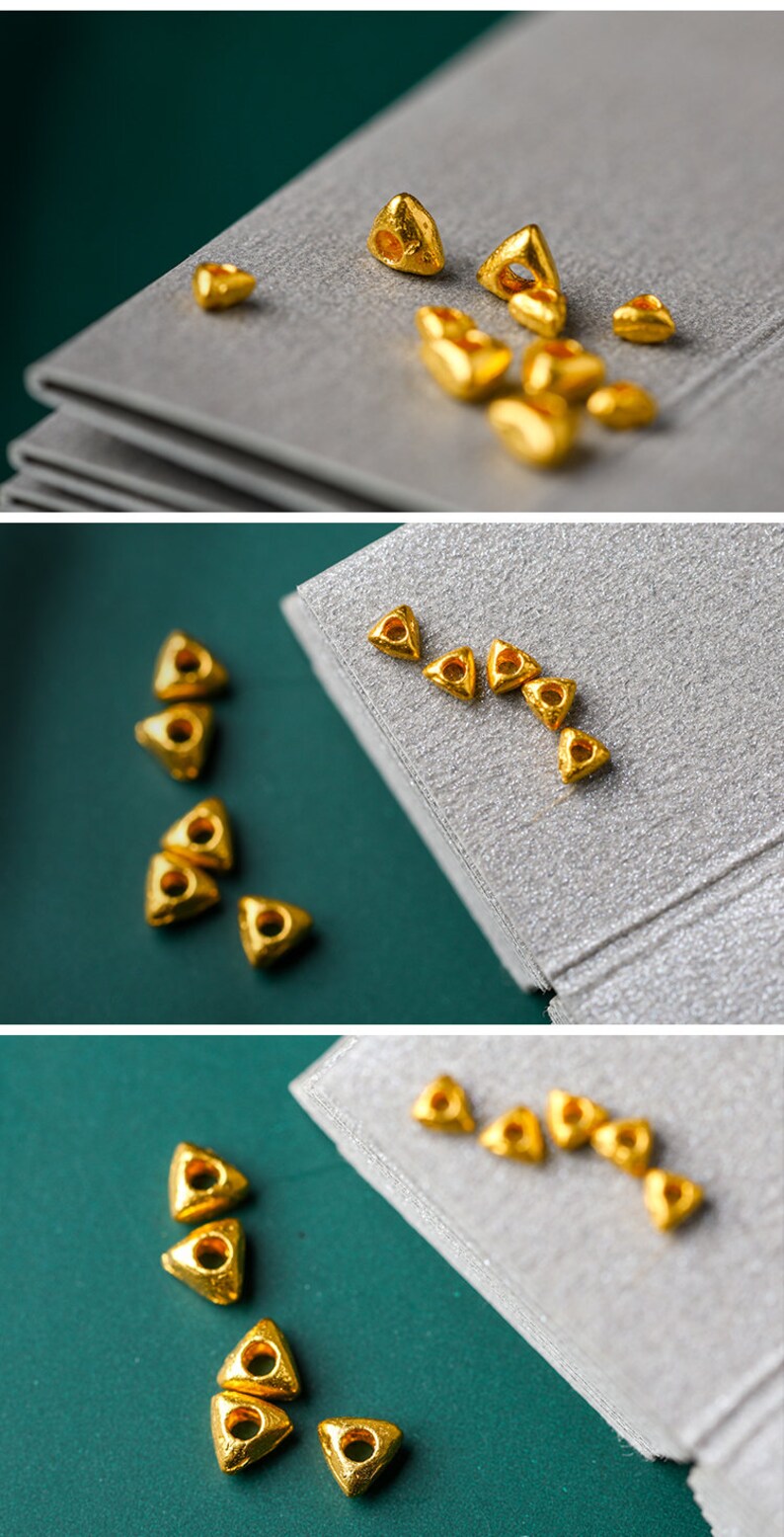May include: A collection of small, gold-colored triangle-shaped beads with a hole in the center. The beads are scattered on a gray surface.