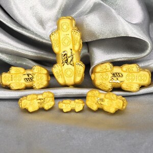 May include: Five gold-colored charms in the shape of a crouching animal, each with intricate details and Chinese characters. The charms are arranged on a silver-colored fabric background.