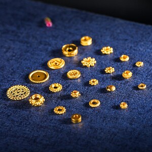 May include: A collection of gold-colored metal beads with various designs and sizes. The beads are arranged on a blue surface.