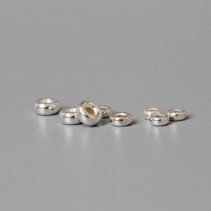 May include: A group of seven silver-coloured metal beads, each with a round, slightly flattened shape. The beads are arranged in a semi-circle on a grey surface.