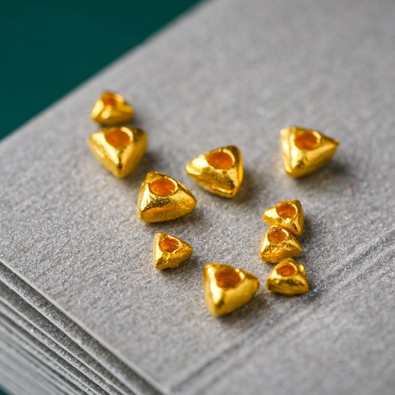 May include: A collection of small, gold, triangular beads with a hole in the center. The beads are scattered on a gray, textured surface.