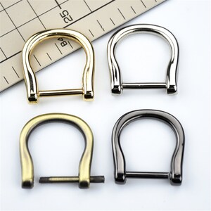 Könnte beinhalten: Vier hufeisenförmige Metallschnallen in Gold, Silber, Antikmessing und Gunmetal-Finish. Die Schnallen haben eine gebogene Oberseite und einen geraden Steg unten mit einer Schraube. Für Lederwaren oder Taschenherstellung.