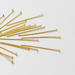 May include: A close-up of a group of gold head pins, also known as jewelry making head pins, scattered on a white surface. The head pins are arranged in a fan shape, with the heads of the pins facing outward.