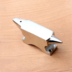 Op de afbeelding: Een kleine, zilverkleurige metalen aambeeld met een puntig uiteinde en een klein gaatje bovenop. Het aambeeld ligt op een houten oppervlak.
