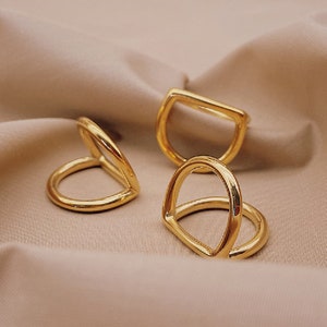 Puede incluir: Tres anillos de metal dorado con diferentes formas. Los anillos están sobre un fondo de tela marrón claro.