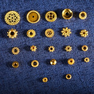 May include: Assortment of gold-colored metal beads and findings, including round, star-shaped, and decorative beads. The beads are arranged in rows on a blue surface.
