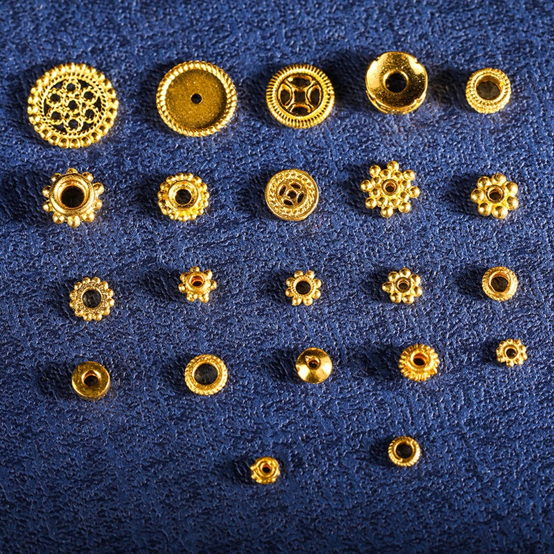 May include: Assortment of gold-colored metal beads and findings, including round, star-shaped, and decorative beads. The beads are arranged in rows on a blue surface.