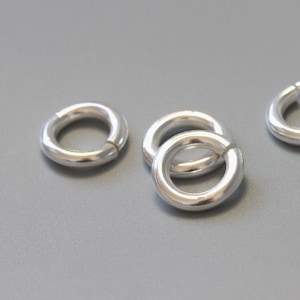 Puede incluir: Cuatro anillos de salto de plata, cada uno con una forma circular y un acabado liso y pulido. Los anillos están dispuestos en un grupo suelto sobre una superficie gris.