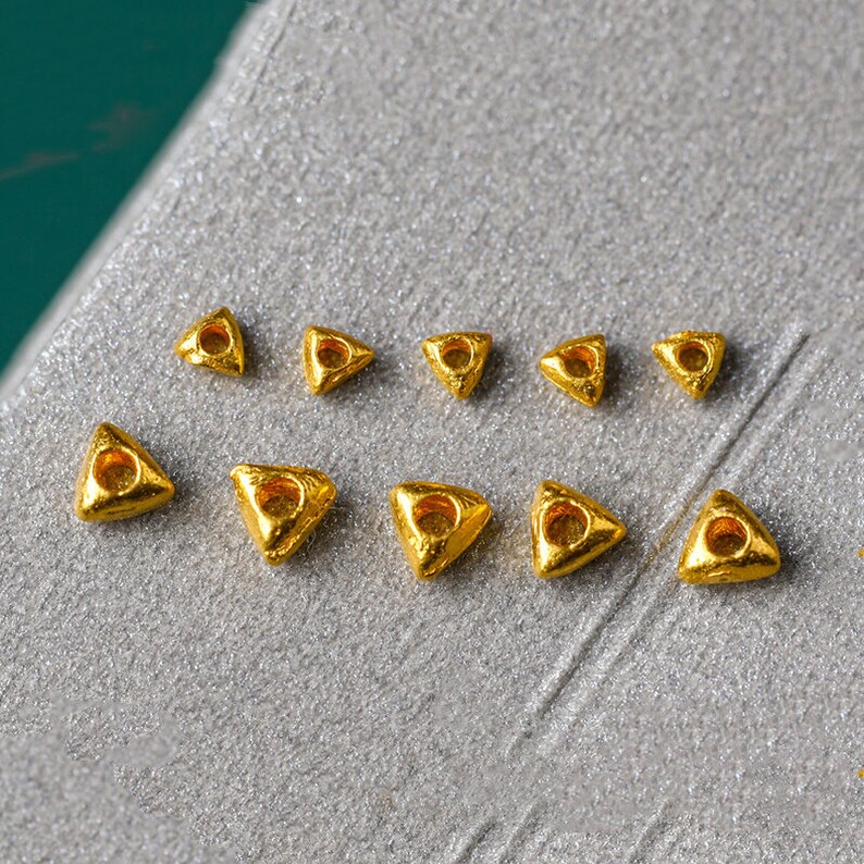 May include: Ten gold triangle-shaped beads with a hole in the center. The beads are arranged in two rows on a silver surface.