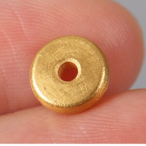 May include: A gold-colored metal bead with a round hole in the center. The bead has a smooth, slightly textured surface.