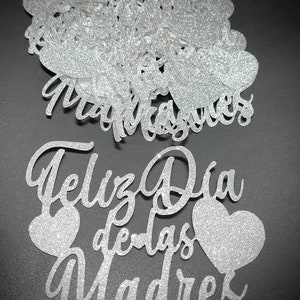 Pu&ograve; includere: Ritagli di carta glitterata argentata con le parole "Feliz Dia de las Madres" e forme di cuore. I ritagli sono in un carattere corsivo e sono sparsi su una superficie scura.
