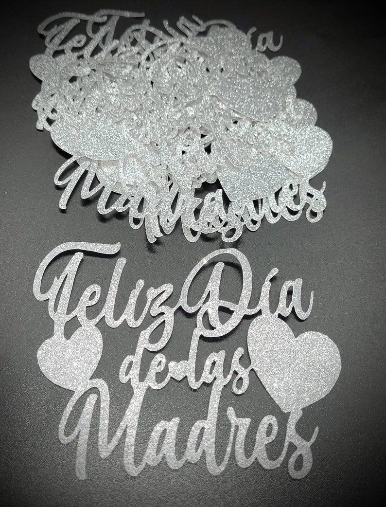 Pu&ograve; includere: Ritagli di carta glitterata argentata con le parole "Feliz Dia de las Madres" e forme di cuore. I ritagli sono in un carattere corsivo e sono sparsi su una superficie scura.