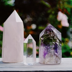 May include: Three crystal points, one pink, one clear, and one purple, are arranged on a white wooden surface. The crystals are all pointed and have a smooth, polished finish.