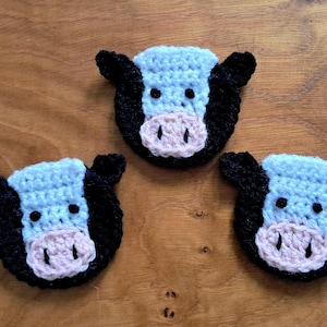 May include: Three handmade crochet cow face appliques. Each cow face features a light blue head, black ears, black eyes, and a pink nose. The appliques are arranged on a wooden surface.