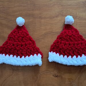 May include: Two handmade red and white Santa hats with white pom-poms. The hats are crocheted with a textured stitch and have a white fluffy trim. Ideal for Christmas decorations or festive crafts.