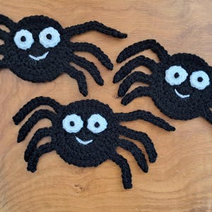 May include: Three black crocheted spider appliques, each featuring a round body with eight spindly legs, two large white circular eyes with black pupils, and a small white stitched smile, crafted from yarn with a textured surface.