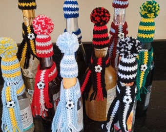 May include: Nine wine bottles with crocheted hats and scarves in various colors and patterns, including black, white, red, blue, yellow, and green. Each hat has a pom-pom on top and the scarves have a soccer ball button.