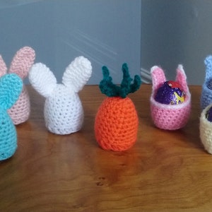 May include: Six crocheted Easter egg holders, each with bunny ears. The holders are in various colours, including pink, white, blue, and orange. Some of the holders have chocolate eggs inside.