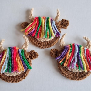 May include: Three handmade crochet Highland cow appliques. Each cow has a brown head, cream horns, and a colourful fringe of yarn in red, yellow, green, and purple. The appliques are set against a white background.