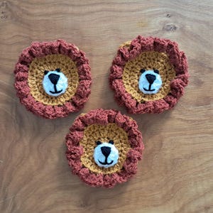 May include: Three handmade crochet lion coasters. Each coaster features a tan centre with a white and black face, surrounded by a rust-coloured mane. The coasters are arranged on a wooden surface.