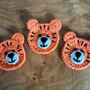 May include: Three handmade crochet tiger face appliques. Each tiger face is orange with black stripes and a light blue and black nose. The appliques are arranged on a wooden surface.