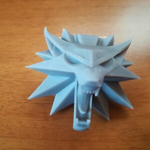 Può includere: Una testa di lupo stampata in 3D di colore azzurro chiaro con un design a stella. La bocca del lupo è aperta e i suoi denti sono visibili.