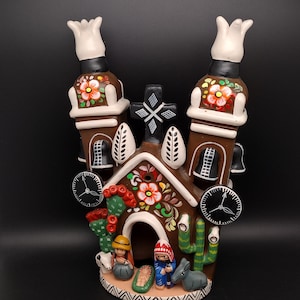 May include: A brown ceramic church-shaped candle holder with white, red, and yellow floral accents. The decorative piece features a cross, clocks, bells, and figures of people and animals. The top has two white tulip-shaped candle holders.