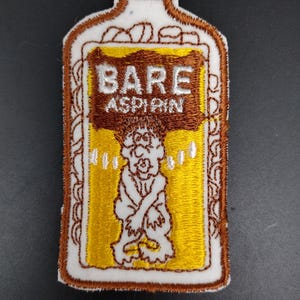 May include: Embroidered patch in the shape of a medicine bottle with the words "BARE ASPIRIN". The patch features a cartoon figure and is outlined in brown with a white and yellow background.