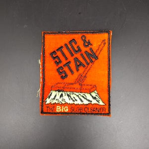 May include: Embroidered orange patch with black text reading "STIC & STAIN" and a broom graphic. The text "THE BIG SLOB CLEANER" is below the broom. The patch has a black border and is likely for clothing or accessories.