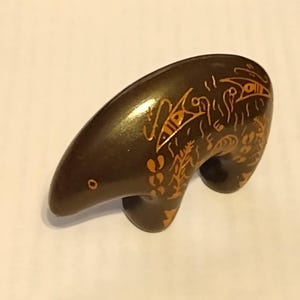 May include: A dark brown, smooth, bear-shaped figurine featuring intricate, light-colored etched patterns along its back and side. The design includes stylized geometric shapes and linear markings that contrast against the dark, polished surface.