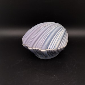 May include: A ceramic seashell-shaped trinket box in shades of lavender, blue, and white. The box has a ridged texture and a glossy finish. The lid fits snugly, and the overall design is reminiscent of a clam shell.