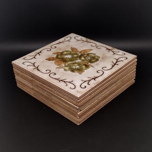 May include: A stack of square ceramic tiles with a cream-colored surface. The top tile features a central floral design in shades of olive green and brown, surrounded by a dark brown decorative vine pattern along the edges of the tile surface.