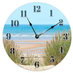 May include: Round clock with a beach scene. The clock face has black numbers on a white background. The background image shows a sandy beach with blue water and green grass.