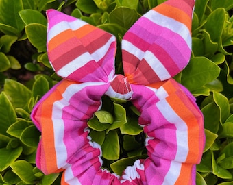 May include: A pink, white, and orange striped scrunchie with a bow on top. The scrunchie is made of fabric and is a popular hair accessory.