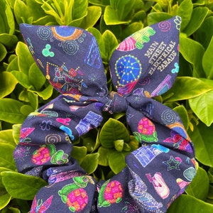 May include: A black scrunchie with a colorful print featuring the text "Main Street Electrical Parade". The print also includes images of fireworks, a carousel, and other carnival-themed elements.