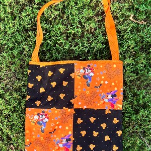 May include: A black and orange Halloween-themed tote bag with a patchwork design featuring Mickey Mouse and Minnie Mouse characters. The bag has a long orange strap.