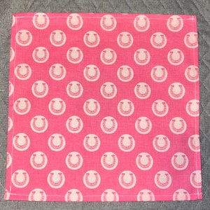May include: Pink fabric with a white horseshoe pattern. The fabric is a square shape and has a white border.