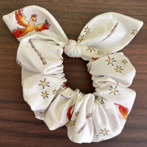 May include: A white fabric hair scrunchie featuring a knotted bow detail. The pattern consists of orange and red phoenix birds, brown wooden wands, and yellow four-pointed stars scattered across the material. The accessory has a gathered, elasticized design.