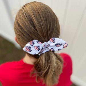 May include: A white hair tie with a red, white, and blue "Vote" design. The hair tie is tied in a bow around a ponytail.