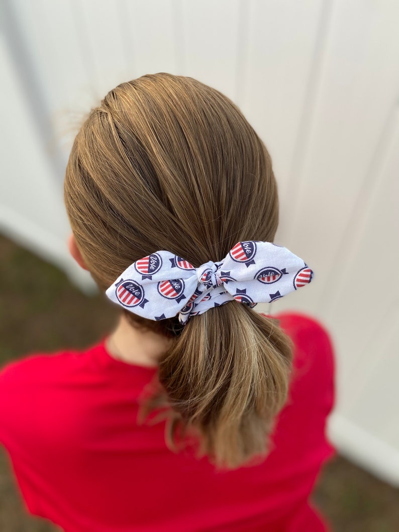 May include: A white hair tie with a red, white, and blue "Vote" design. The hair tie is tied in a bow around a ponytail.