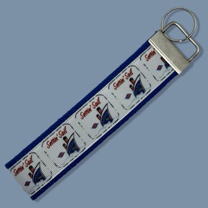 May include: A blue and white key chain with a white background and a blue border. The key chain has a repeating pattern of a ship with the text "Settin' Sail" on it. The key chain has a silver key ring.