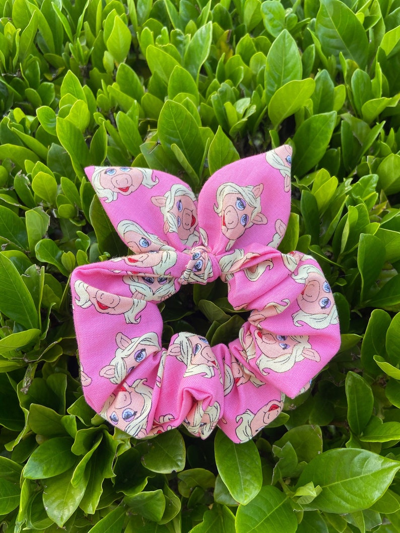 Peut inclure: Un chouchou rose avec un motif r&eacute;p&eacute;t&eacute; du visage de Miss Piggy.