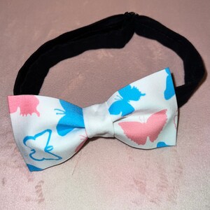 May include: White bow tie with a black adjustable strap. The bow tie features a pattern of pink and blue butterflies.