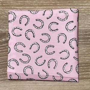 May include: A square, pink fabric item with a repeating pattern of silver horseshoes. The horseshoes are detailed with a dark outline and small dot accents. The fabric has a soft, slightly textured appearance.
